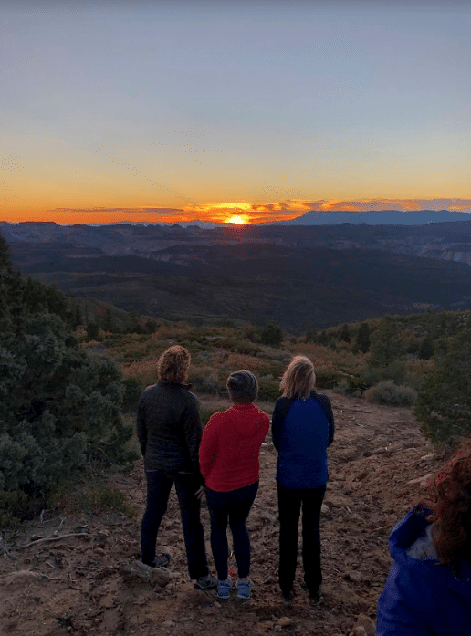 Three people stand on a hillside, gazing at a sunset over a mountainous landscape.